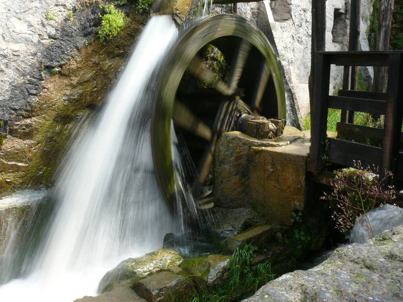 Una ruota idraulica in legno e ferro mossa da un salto d'acqua, incassata fra la roccia e la vegetazione.