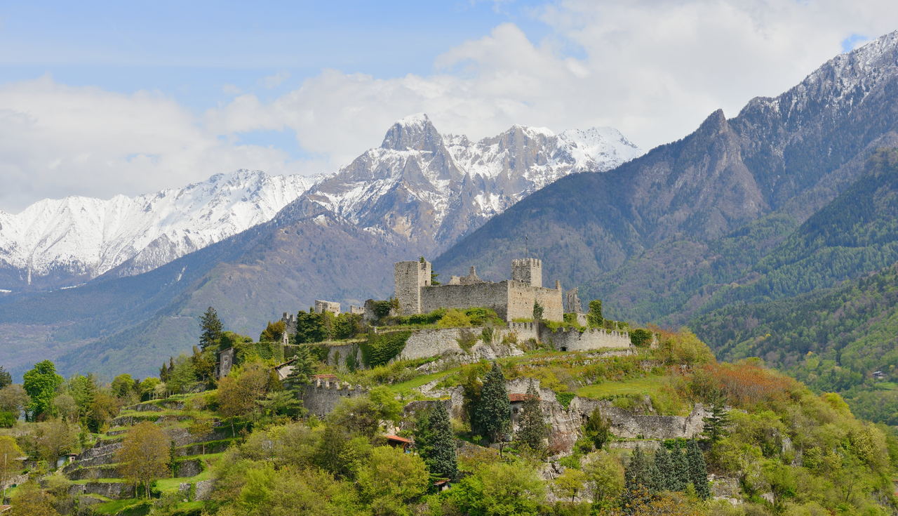 Il castello di Breno sulla sommità del colle, con le mura e le torri in pietra sopra il verde e le montagne innevate alle spalle.