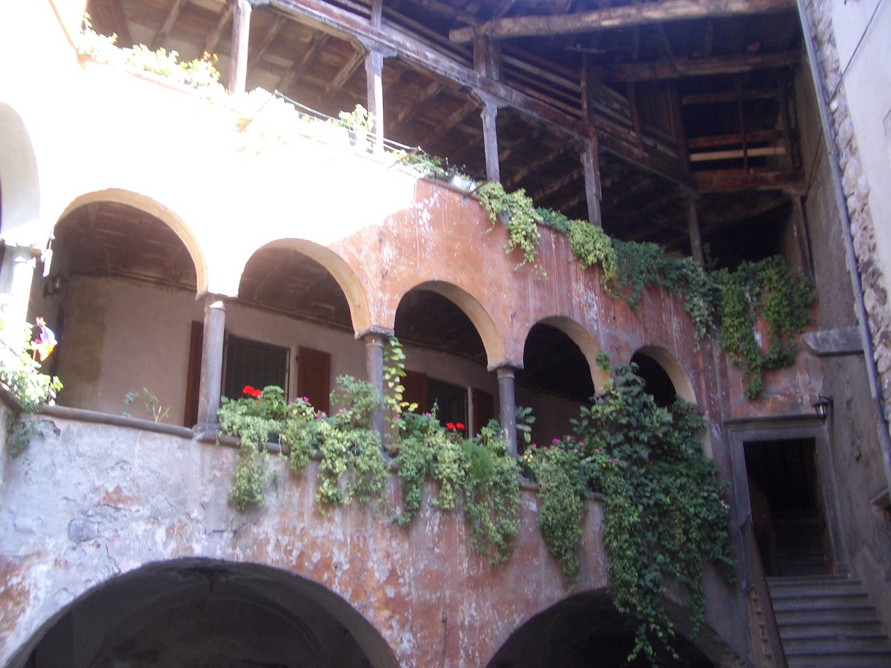 Loggia in pietra a cinque arcate su colonne, con ballatoio in legno al piano superiore, gerani alle aperture e una scala in pietra sulla destra.