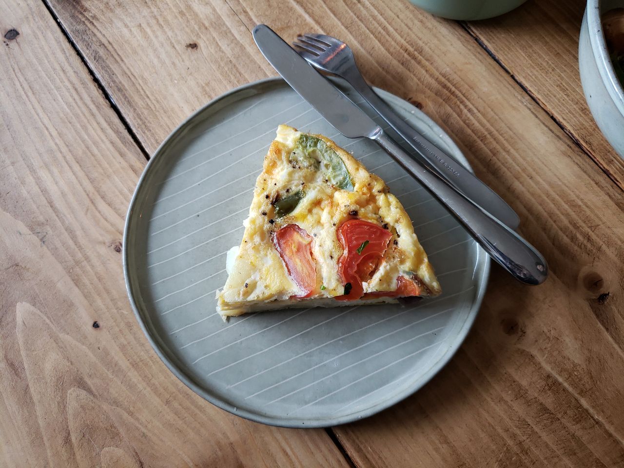 Una fetta di frittata servita nel piatto, con pomodoro e peperone verde, e le posate appoggiate accanto su un tavolo di legno.