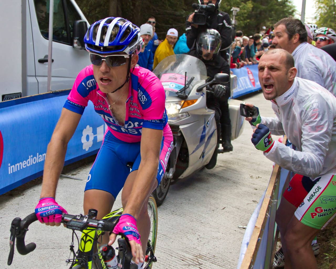 Un corridore in salita sul Mortirolo durante il Giro d'Italia, affiancato dal pubblico a bordo strada e dall'ammiraglia della squadra.