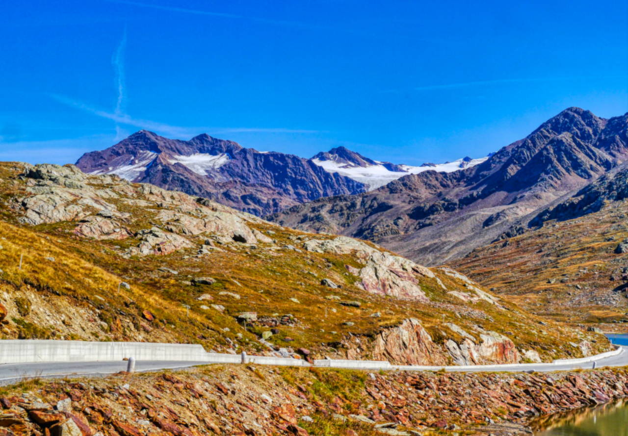 La strada del Passo Gavia in prossimità del valico, fra rocce e praterie d'alta quota, con le cime e i nevai del gruppo sullo sfondo.