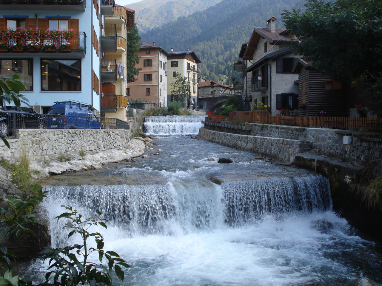 Il corso dell'Oglio incanalato fra le case di Ponte di Legno, con una briglia che increspa l'acqua e gli edifici del centro sulle sponde.
