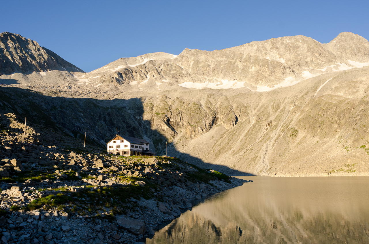Il rifugio Garibaldi in muratura sulle rive di un lago d'alta quota, circondato da pietraie e dalle cime dell'alta Val d'Avio.