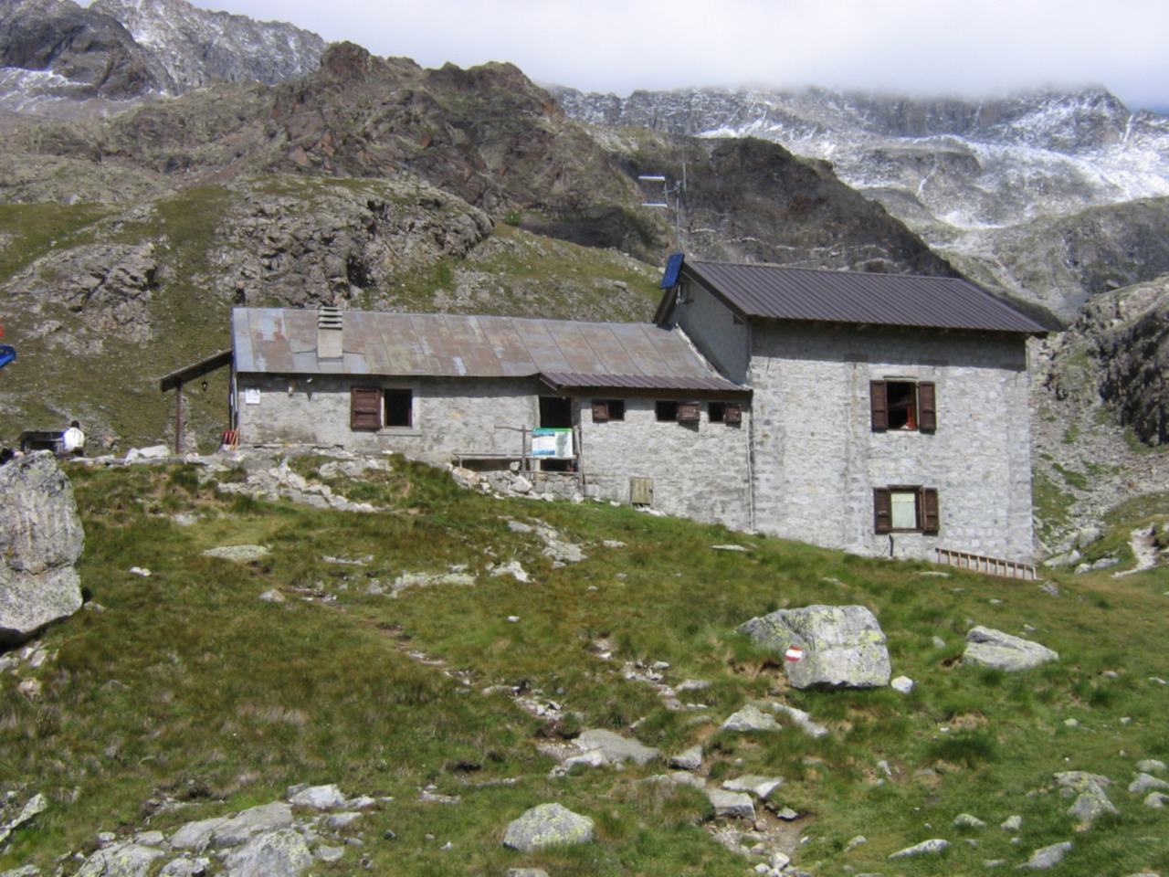Un rifugio in pietra su un dosso d'alta quota, sotto creste rocciose ancora innevate e nuvole basse; su un masso in primo piano il segnavia bianco-rosso.