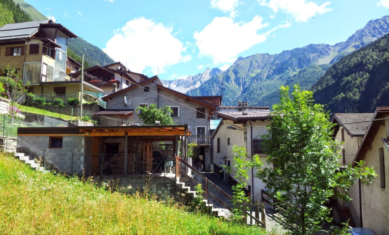 Una via di Saviore dell'Adamello fra case in pietra con balconi in legno, e i versanti boscosi della valle sullo sfondo.
