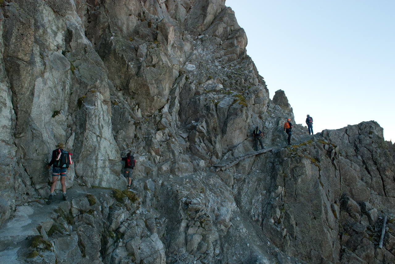 Escursionisti in fila su una cengia attrezzata con cavo metallico, sospesa su una parete rocciosa in alta quota.