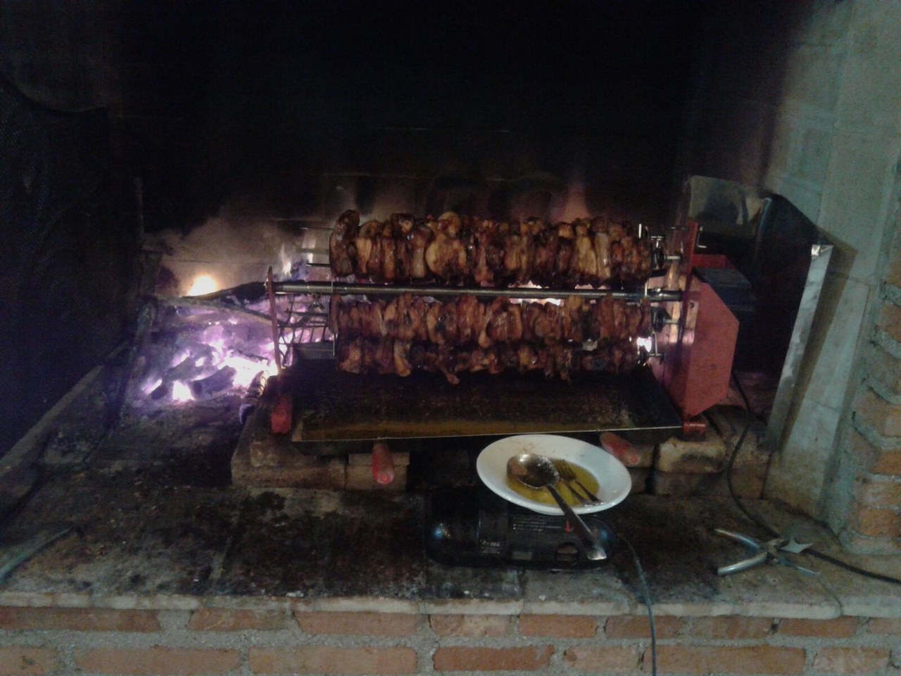 Due lunghi spiedi di carne che girano sulla brace dentro un camino in mattoni, con il motore dello spiedo sulla destra.